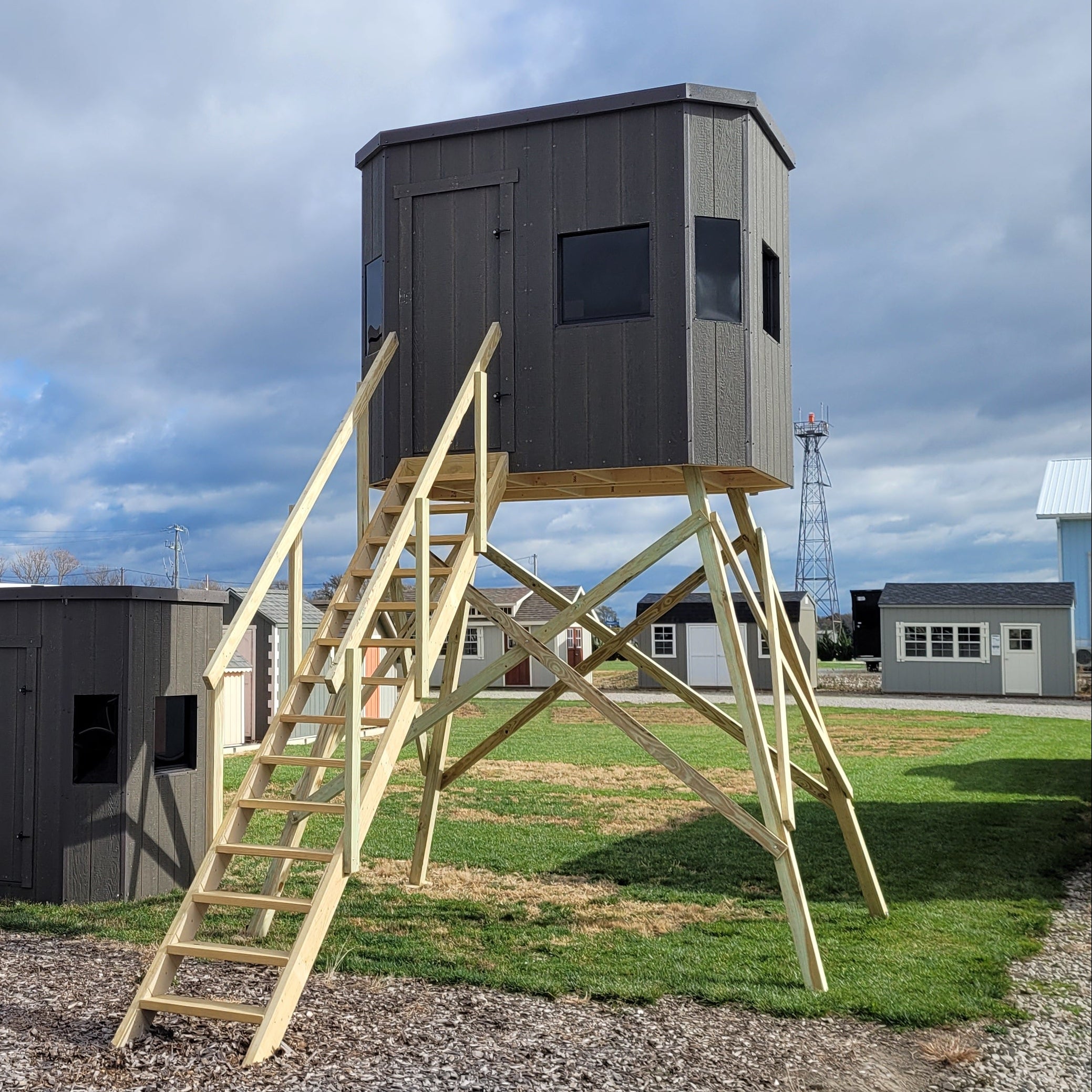 Gray elevated hunting blind stand on wooden A-frame legs with attached wooden stairs, shown outdoors on a gravel and grass lot under a cloudy sky.