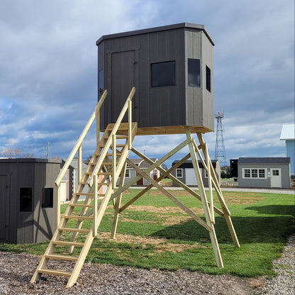 Gray elevated hunting blind stand on wooden A-frame legs with attached wooden stairs, shown outdoors on a gravel and grass lot under a cloudy sky.