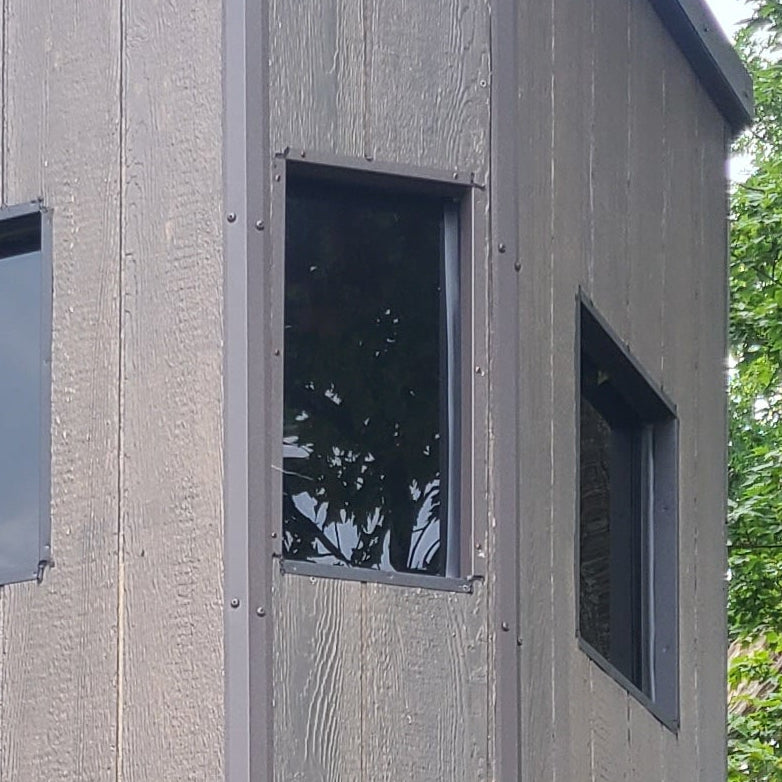 A wooden elevated hunting stand with a gray cabin structure and a tall natural wood support frame, photographed outdoors among trees and grass.