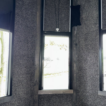 A hunting blind interior corner with three tall windows set in carpeted gray walls, showing a bright outdoor scene with trees through the glass.